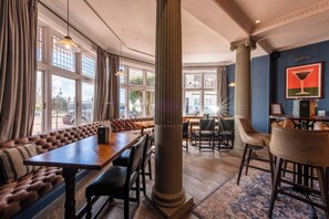 A restaurant interior with a long bench seating area, a high ceiling, and large windows.