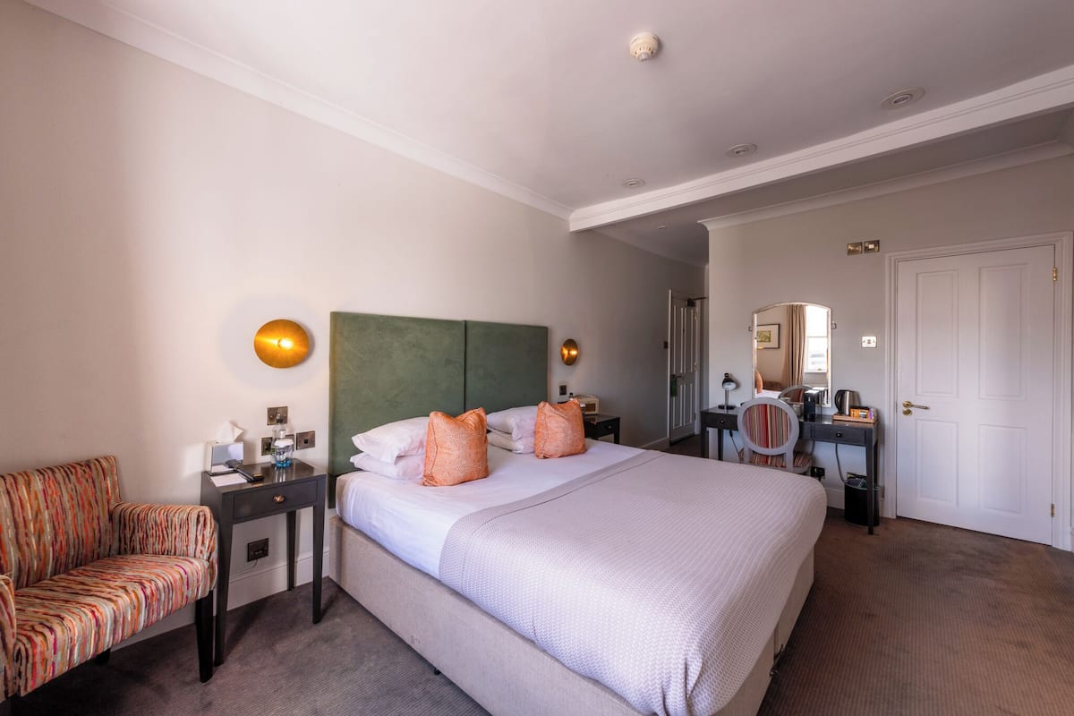 A hotel room with a large bed, a green headboard, a bedside table with a lamp, a chair, and a desk with a mirror and a small table.