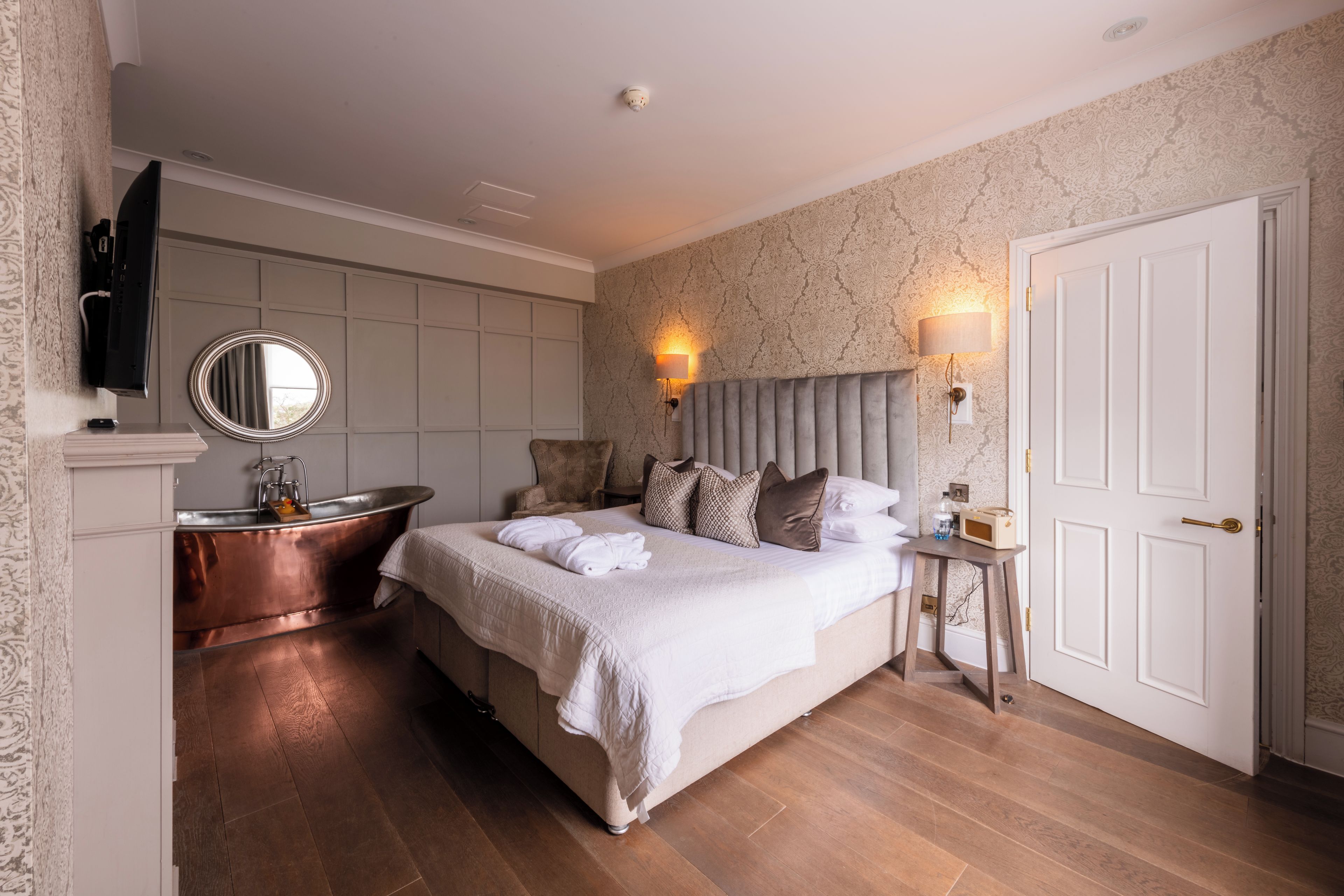 A hotel room with a large bed, a sitting area with a chair, a vanity with a mirror, a bathtub, and a television mounted on the wall.