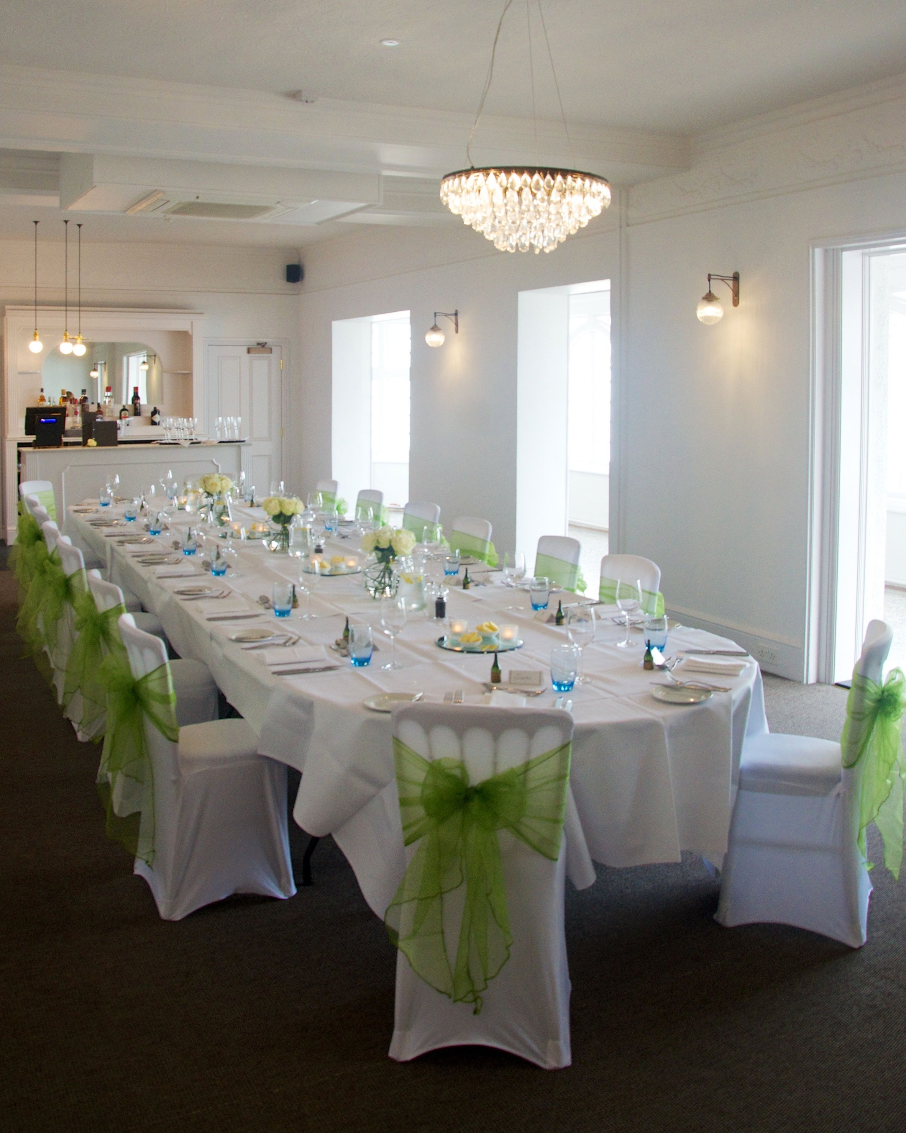 A long banquet table set for a formal event, with chairs adorned with green sashes, white tablecloths, and a chandelier overhead.