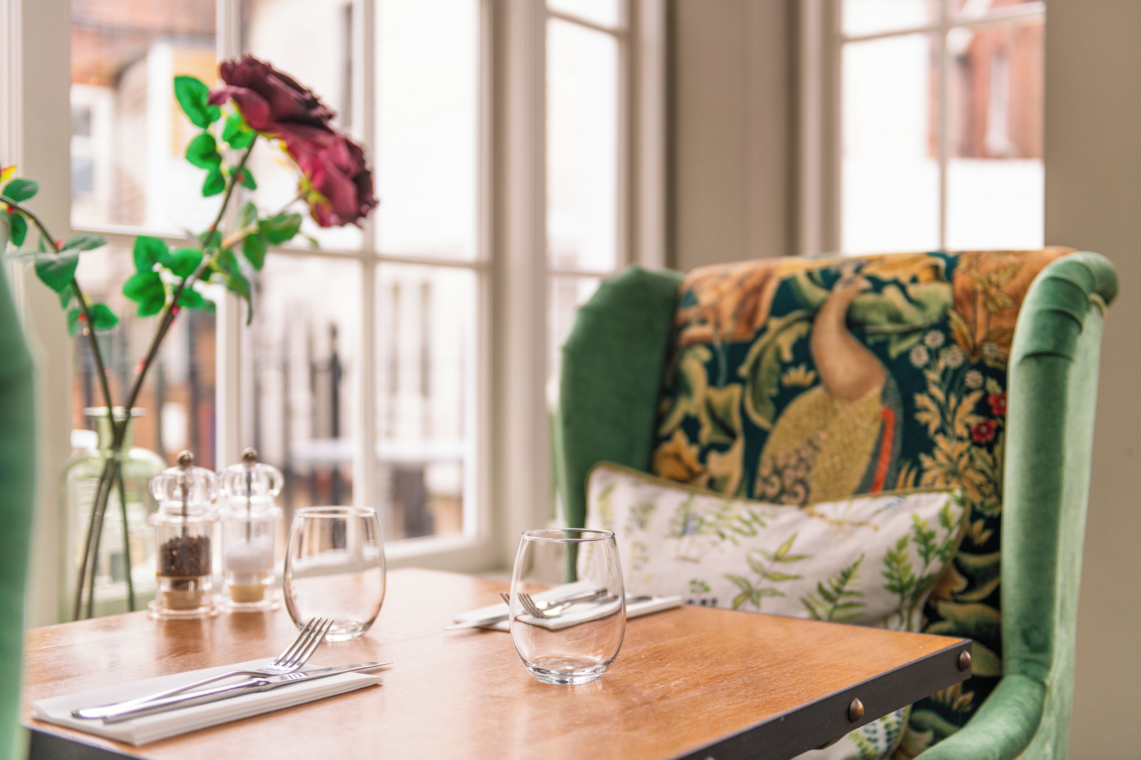 Una mesa de comedor con una silla tapizada de verde, cristalería transparente y un florero con una flor.