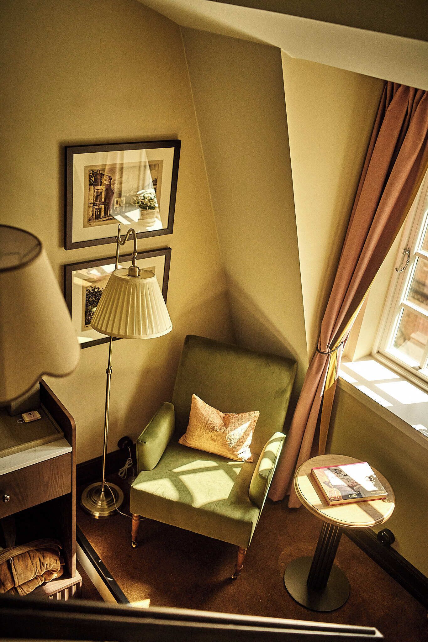A corner room with a green armchair, a standing lamp, a framed picture on the wall, and a window with curtains.