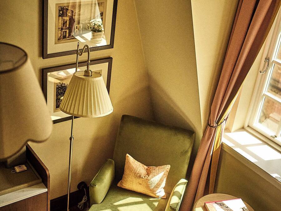 A corner room with a green armchair, a standing lamp, a framed picture on the wall, and a window with curtains.