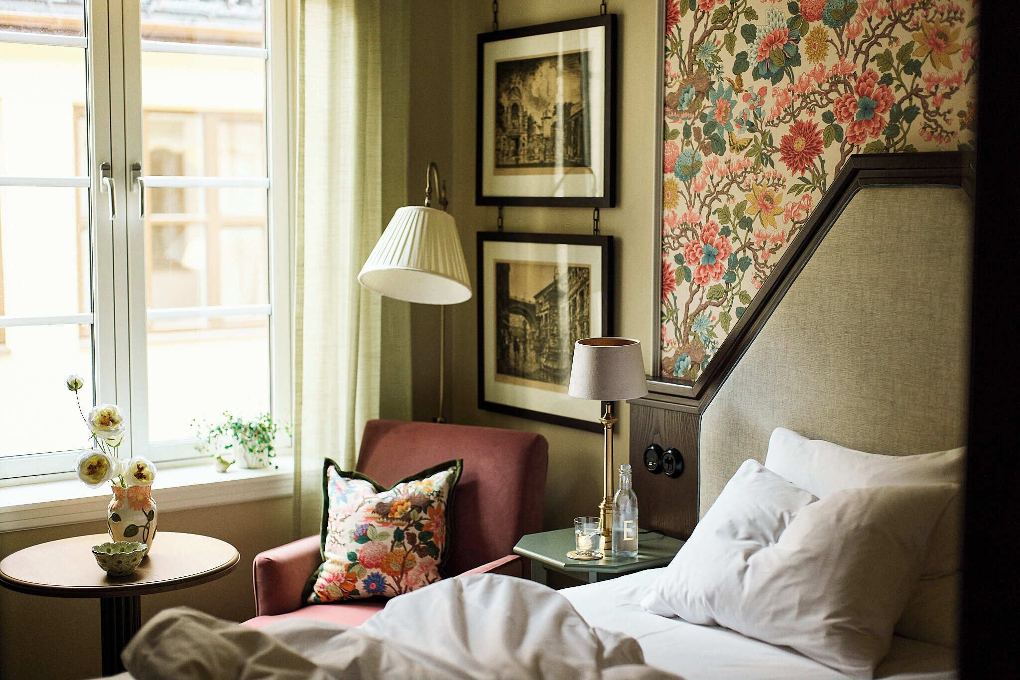 Habitación de hotel con cama, una silla, una mesita de noche con lámpara, un jarrón con flores y una ventana con vistas.