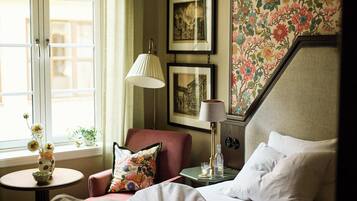 Habitación de hotel con cama, una silla, una mesita de noche con lámpara, un jarrón con flores y una ventana con vistas.