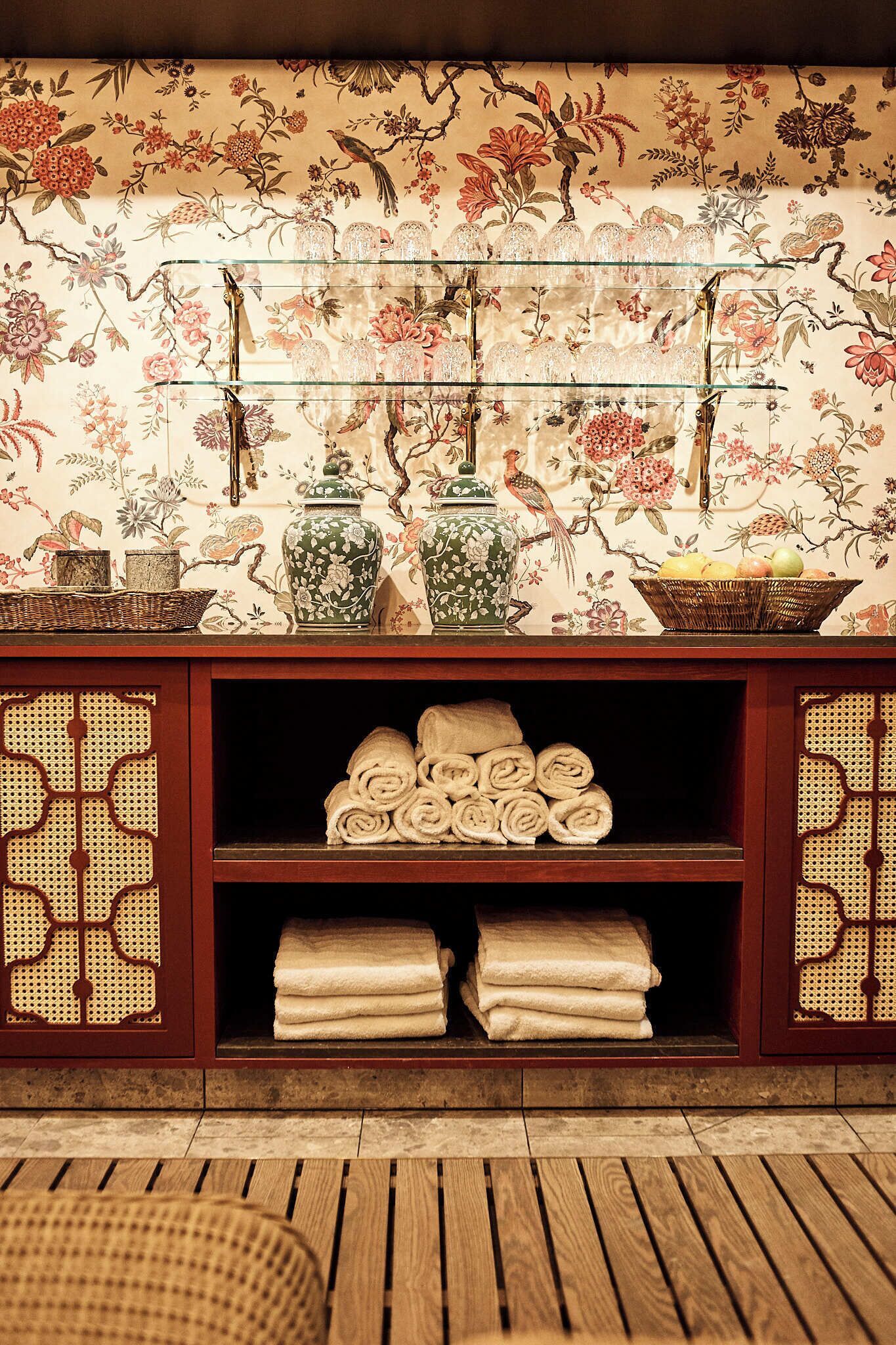 A room with a wooden floor, a decorative wall with floral patterns, a glass shelf with jars and a basket, and a shelf with neatly folded towels.