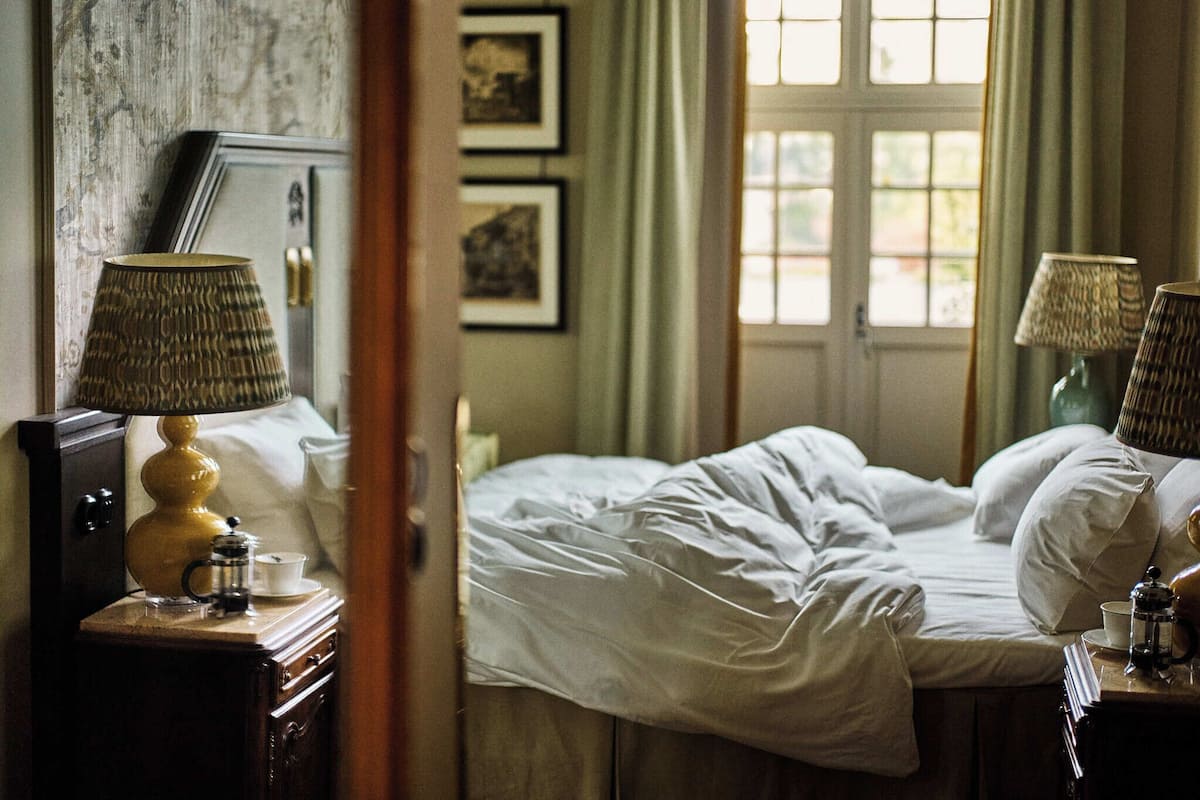 Une chambre avec un lit, deux tables de chevet avec des lampes, une fenêtre avec des rideaux et des tableaux encadrés au mur.
