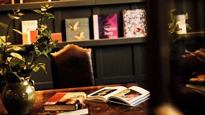 Una habitación con una estantería, una mesa con libros, un florero con una planta y una lámpara colgante.
