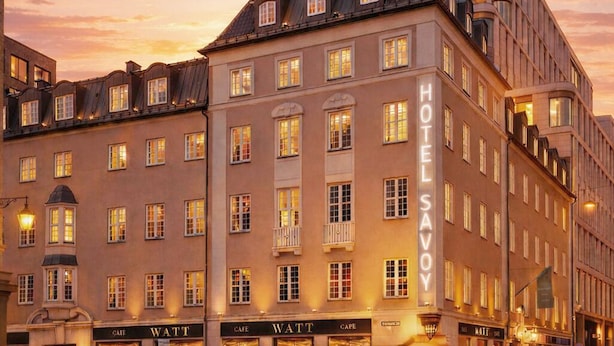 Un hotel de estilo arquitectónico europeo clásico, con un llamativo cartel de 'HOTEL SAVOY', varios locales comerciales a nivel de calle y una fachada bien iluminada.