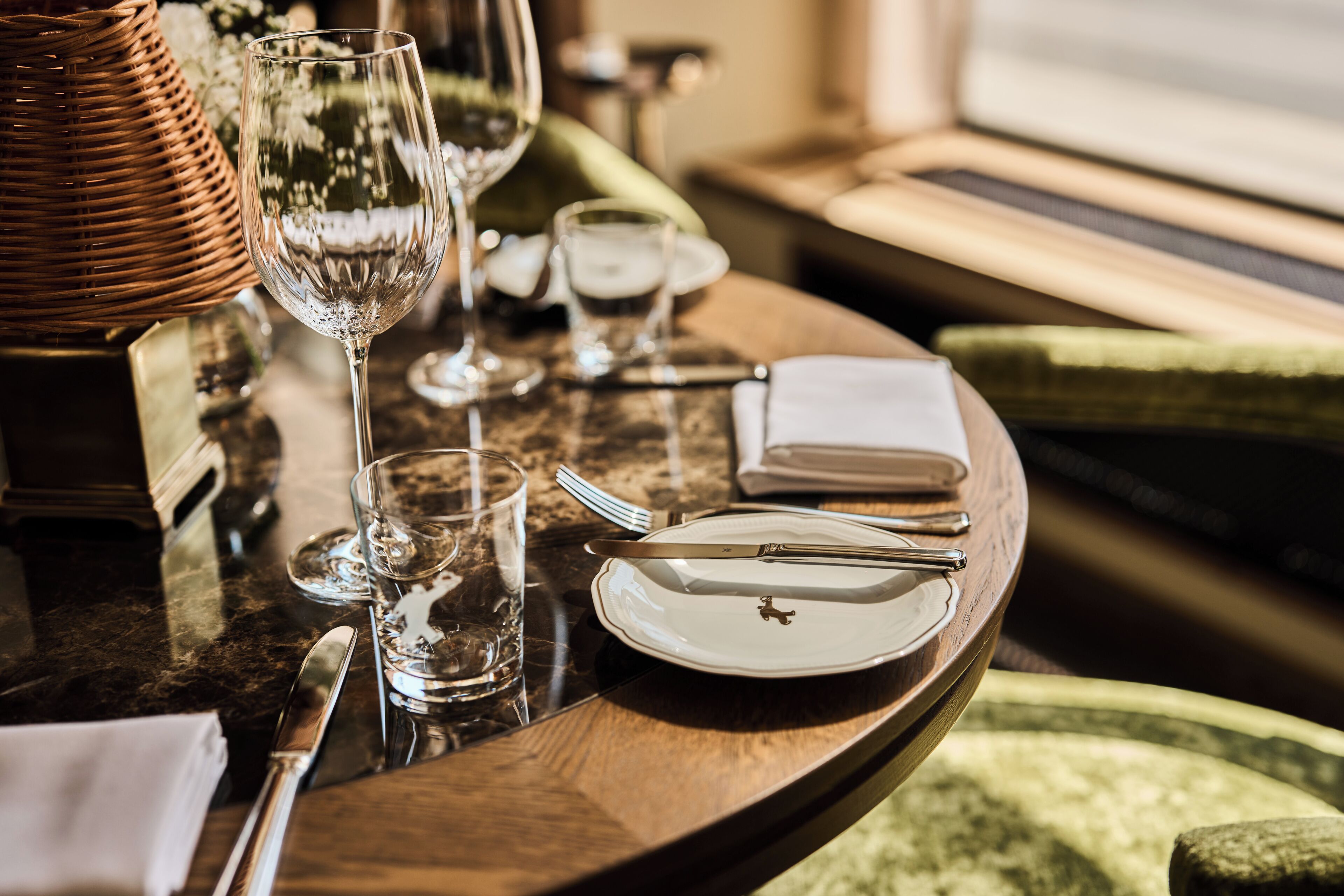 A dining table with a plate, cutlery, and glasses, set in a room with a window and green upholstered furniture.