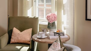 Una habitación acogedora con un sillón verde, una mesita redonda con un jarrón de flores rosas, una lámpara y una ventana con cortinas.