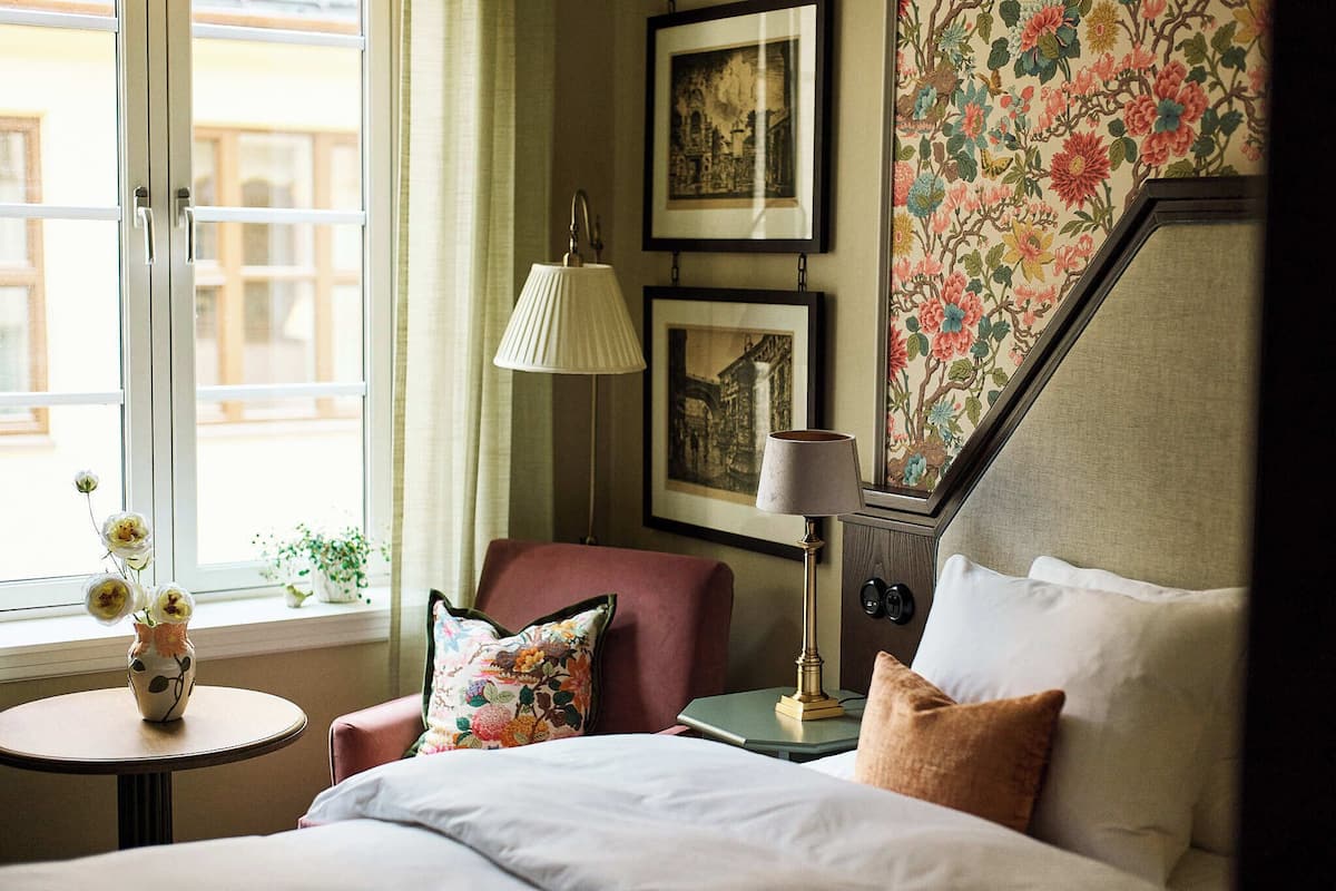 Une chambre d’hôtel avec un lit, une chaise, une petite table sur laquelle se trouve un vase, une lampe et des tableaux encadrés accrochés au mur.