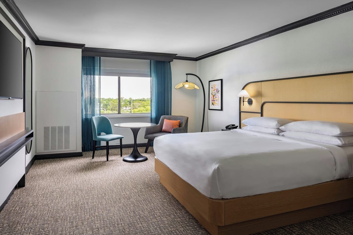 A hotel room with a large bed, a desk, a chair, a window with curtains, and a lamp.