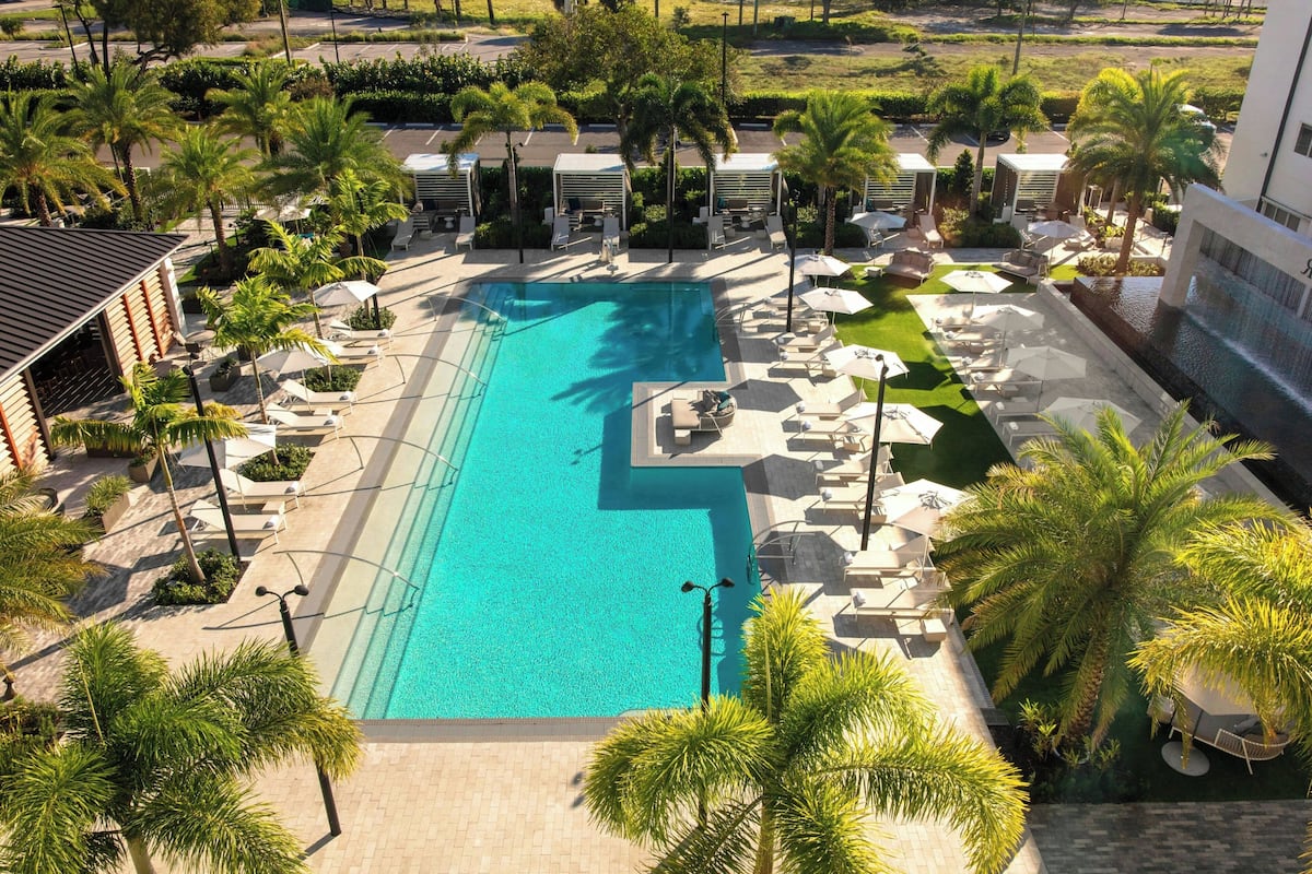 A large swimming pool surrounded by palm trees and lounge chairs, with a building in the background.