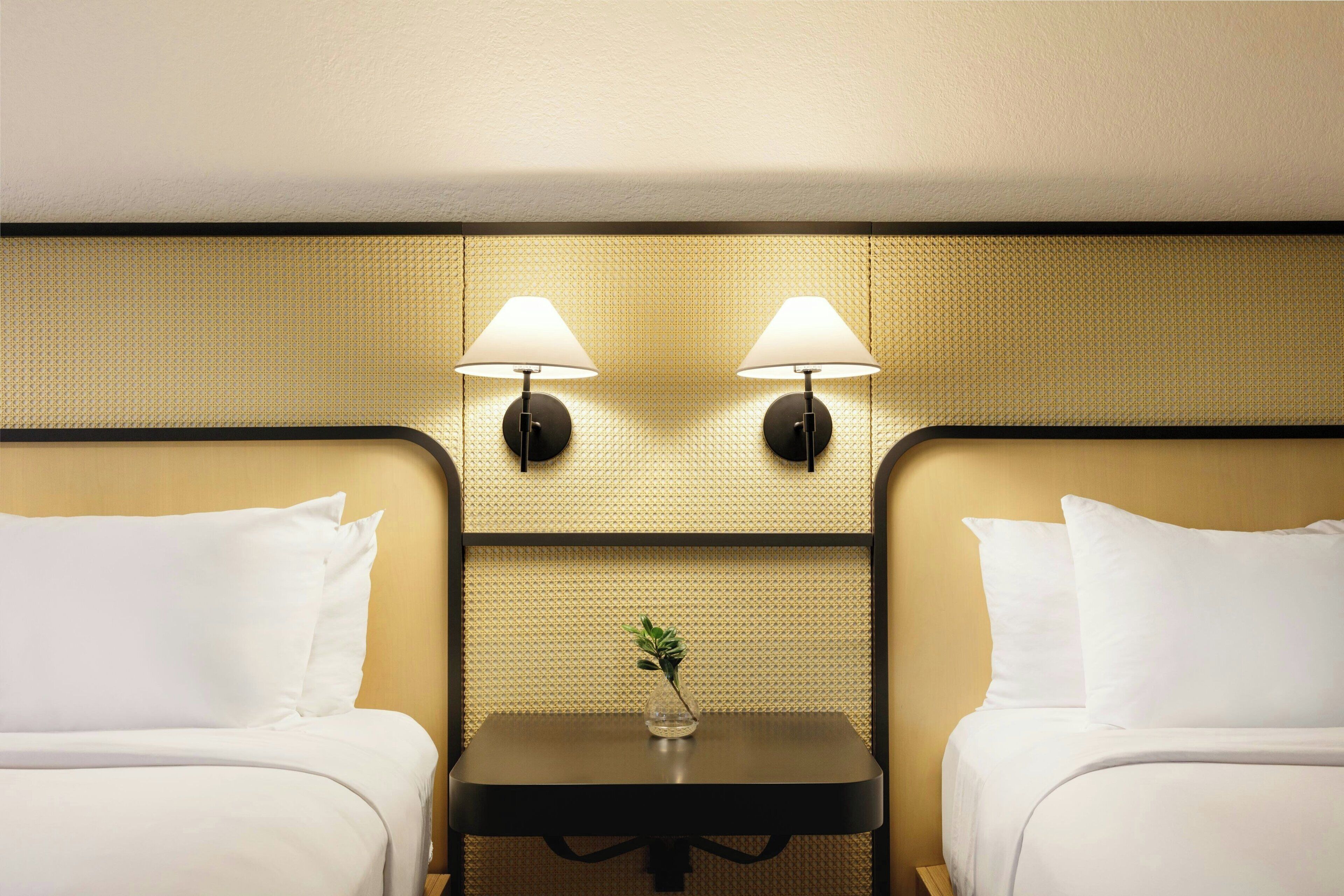A hotel room with two beds, a small table with a vase, and two wall-mounted lamps.