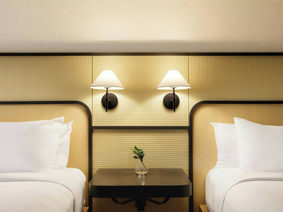 A hotel room with two beds, a small table with a vase, and two wall-mounted lamps.