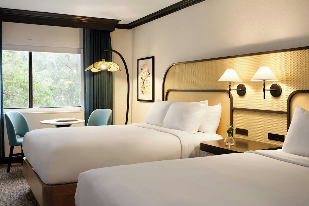 A hotel room with two beds, a small table, a chair, and a window with a view of trees.