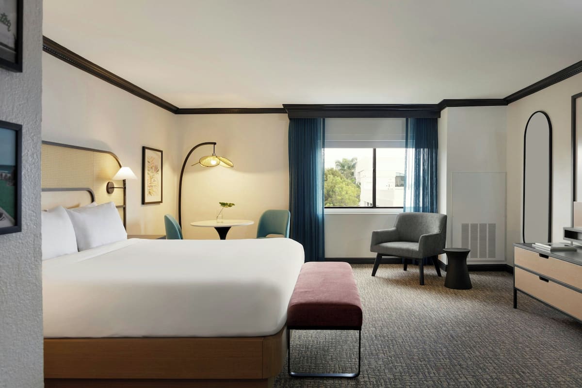 A hotel room with a large bed, a desk, a chair, a small table, and a window with curtains.