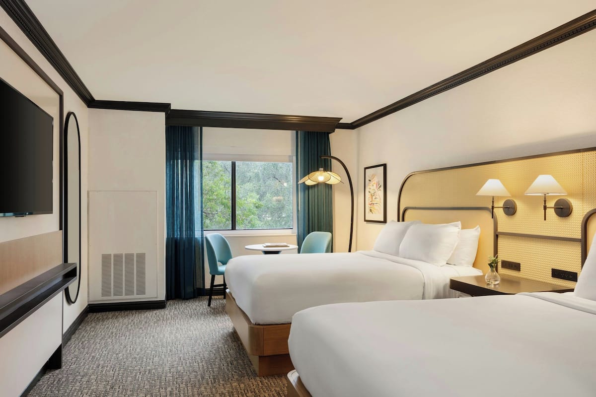 A hotel room with two beds, a TV, a small table, and a window with curtains.