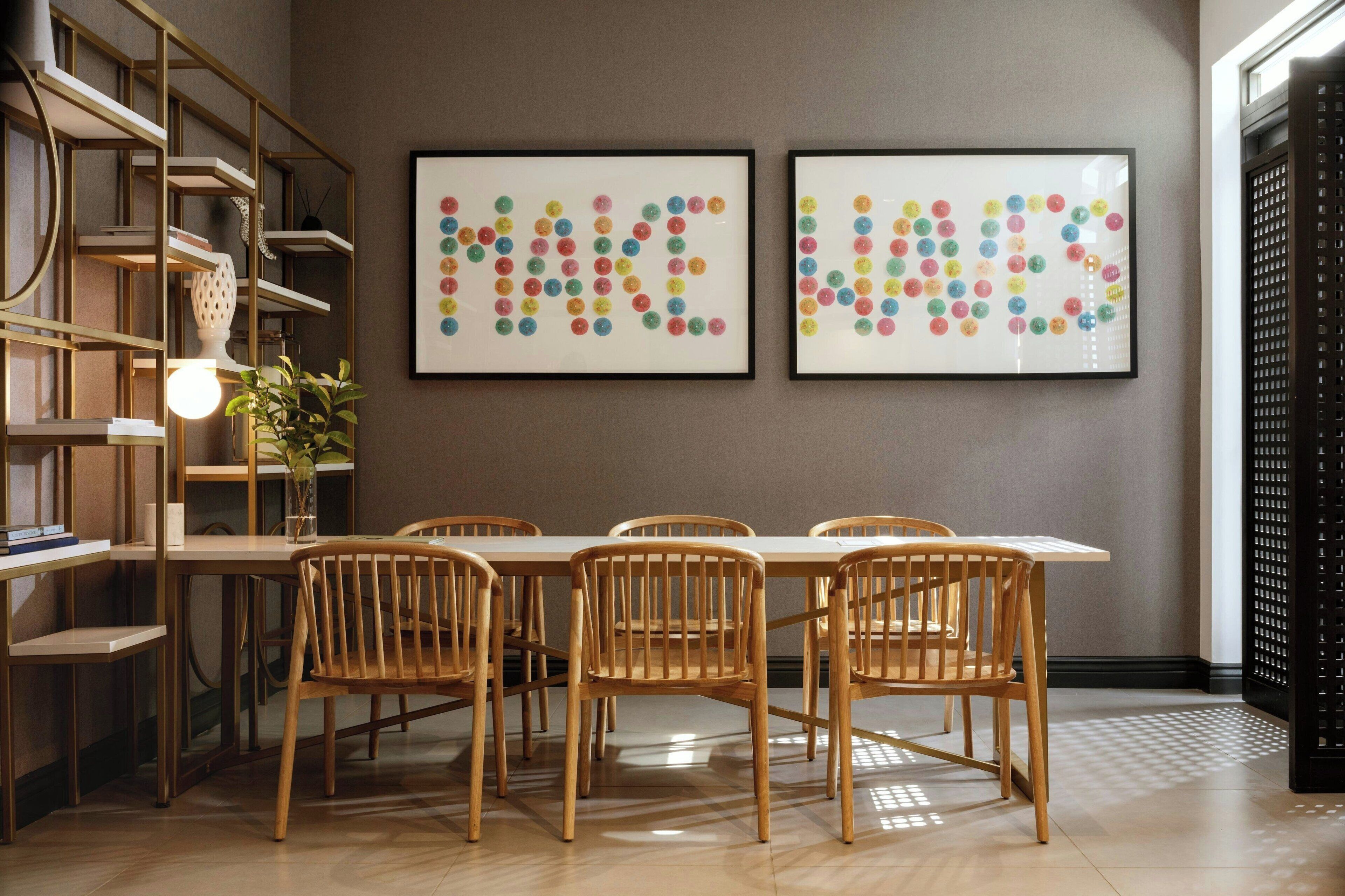 A dining area with a long table, six wooden chairs, and two framed artworks on the wall.