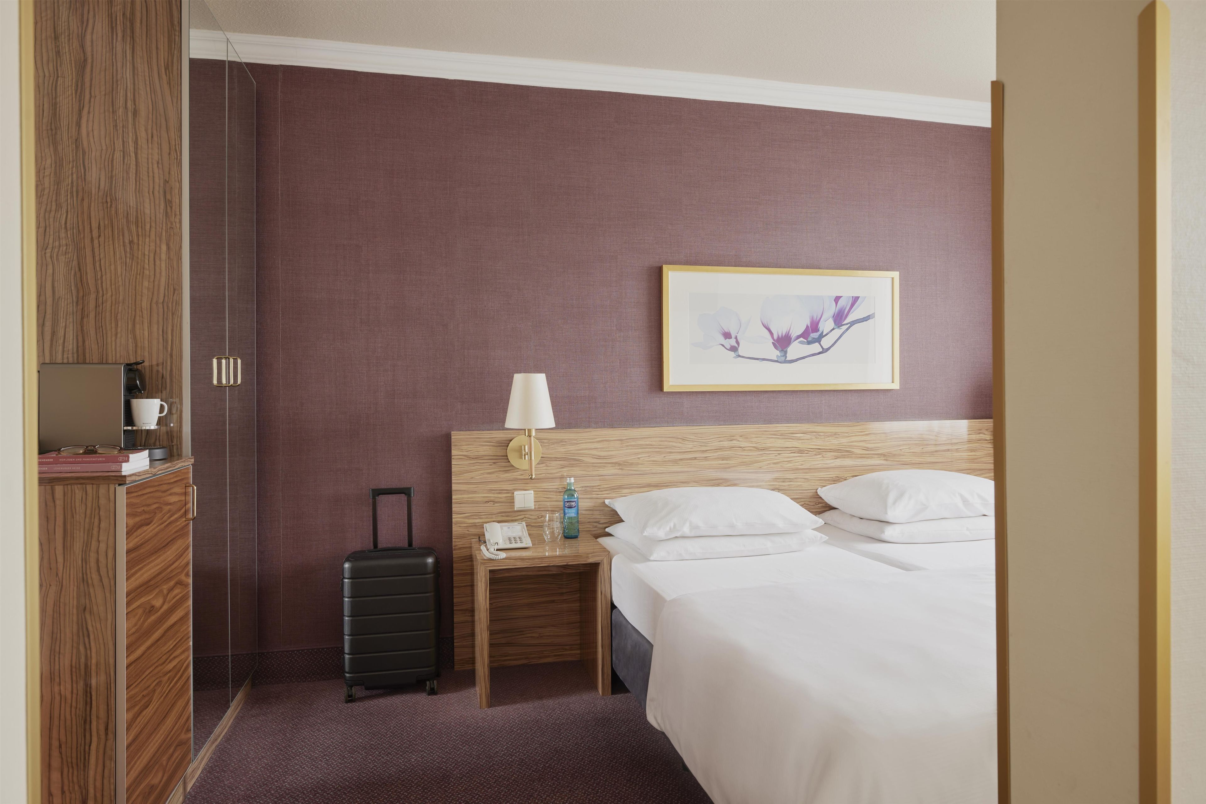 Ein Hotelzimmer mit Bett, Nachttisch, Lampe, einem Bild an der Wand und einem Koffer.