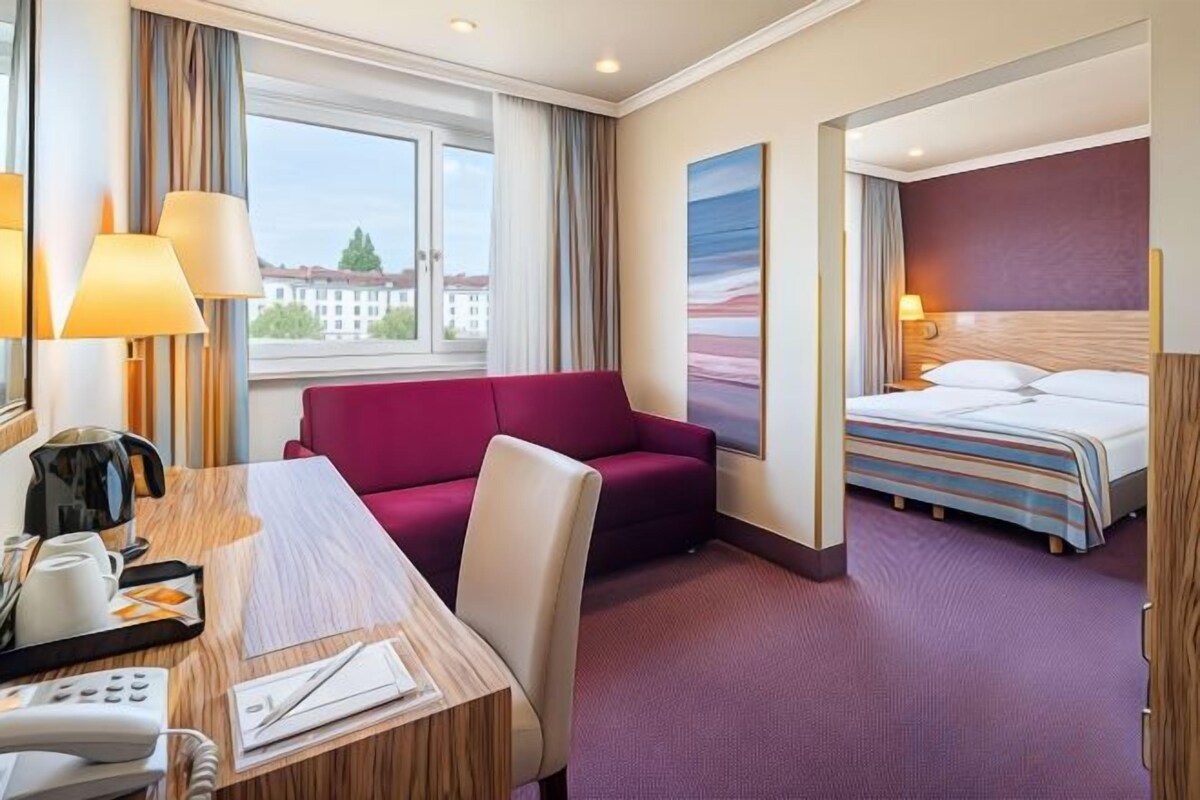 A hotel room with a bed, a desk with a telephone, a chair, a sofa, and a window with curtains.