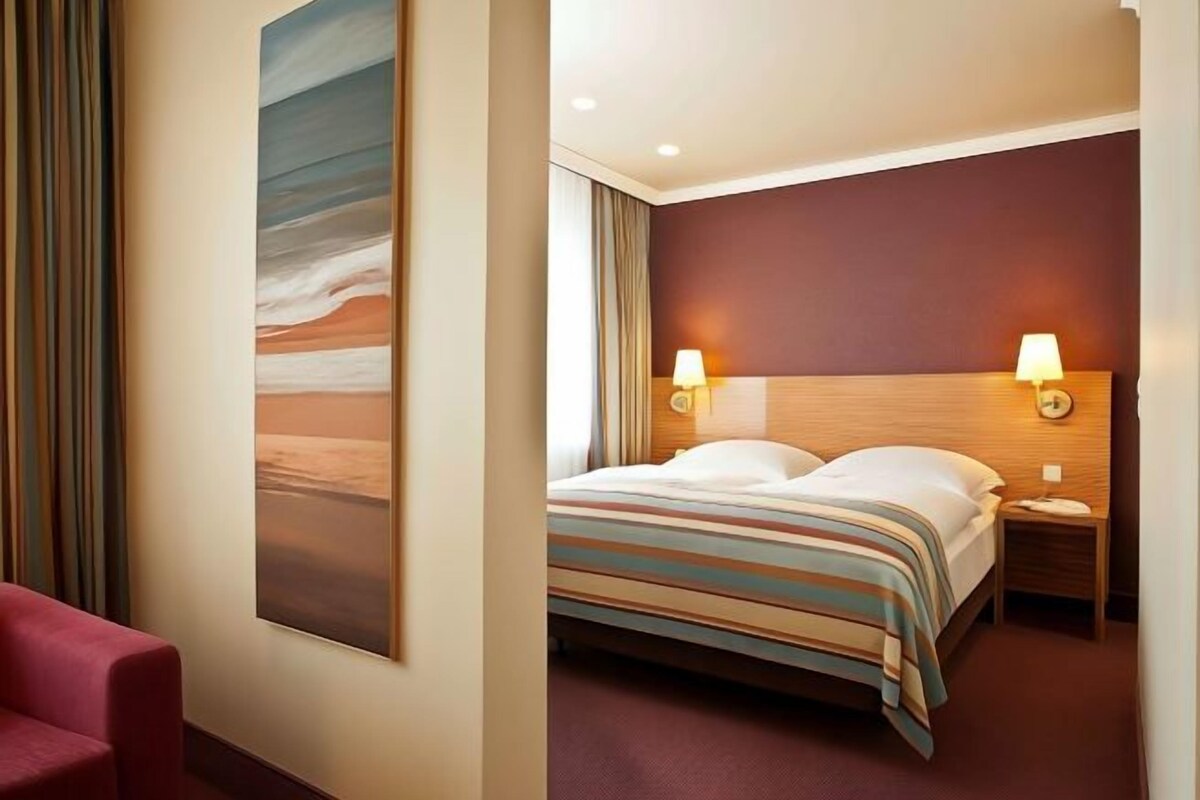 A hotel room with a bed, two bedside lamps, a large painting on the wall, and a red chair.