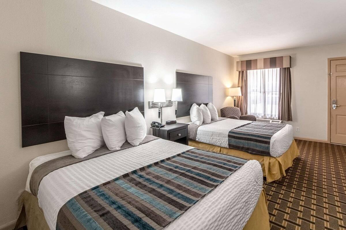 A hotel room with two beds, a nightstand, a lamp, and a window with curtains.