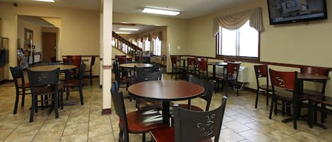 Un grand espace dédié à la salle à manger, avec plusieurs tables et chaises, un téléviseur fixé au mur et un sol carrelé.