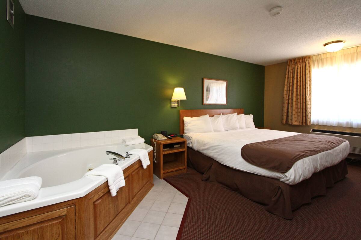 Habitación de hotel con una bañera grande, una cama con colcha marrón, una mesita de noche con lámpara y un cuadro en la pared.