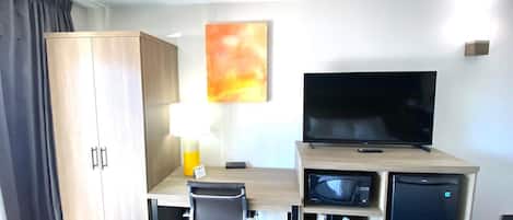 A hotel room with a wooden desk, a television, a microwave, a refrigerator, a chair, and a cabinet.