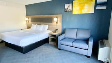 Habitación de hotel con una cama grande, un sillón gris, una mesita de noche con lámpara y una obra de arte en la pared.