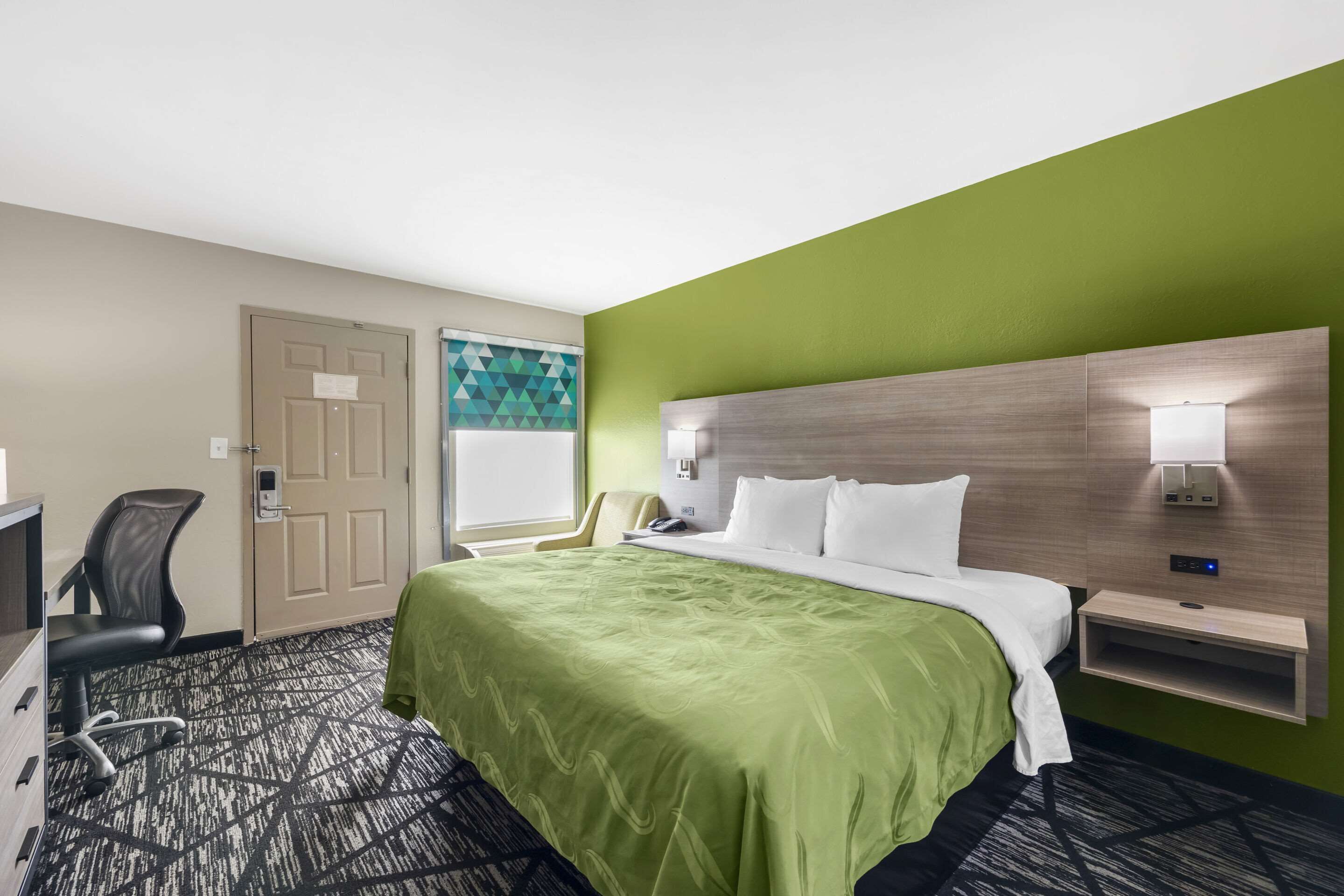 A hotel room with a large bed, a desk, a chair, and a nightstand with a lamp.