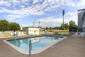 A rectangular swimming pool with a metal ladder, surrounded by a concrete deck. There are white lounge chairs and a small building with a sign in the background.