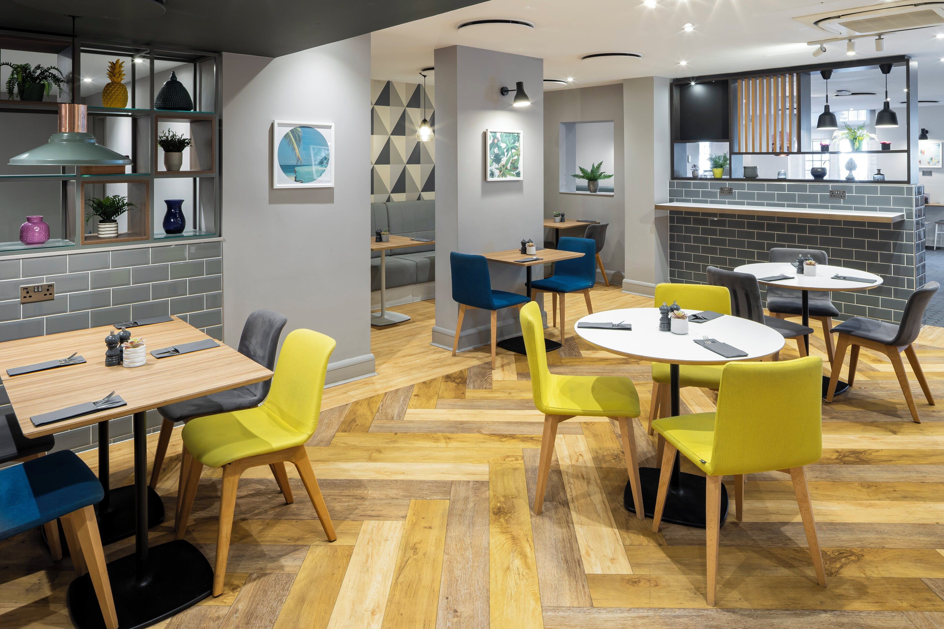 A modern cafe with wooden tables and bright yellow chairs, a bar area with a black countertop, and a herringbone wood floor.