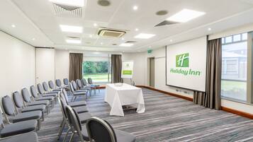 A conference room with rows of chairs, a table with a white tablecloth, a projection screen with the Holiday Inn logo, and a view of greenery outside.