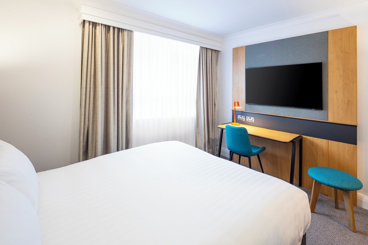A hotel room with a large bed, a desk with a chair, a television mounted on the wall, and a window with curtains.