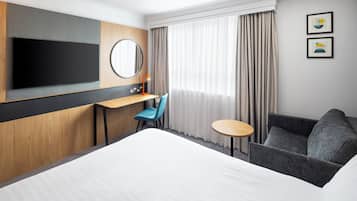 A hotel room with a large bed, a desk with a chair, a small round table, a sofa, a TV mounted on the wall, and two framed pictures on the wall.