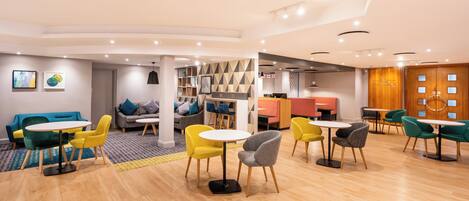 A modern, well-lit lounge area with round tables and colorful chairs, a seating area with sofas, and a reception desk.