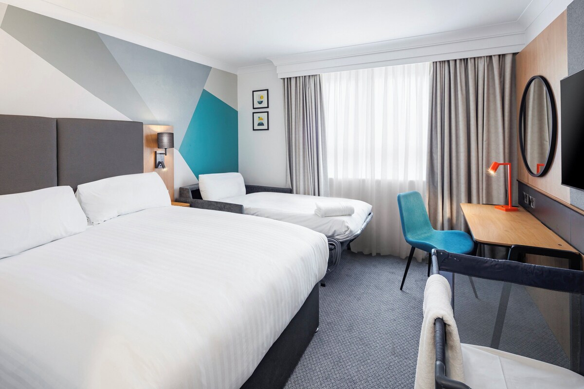 A hotel room with two beds, a desk, a chair, a TV, and a window with curtains.