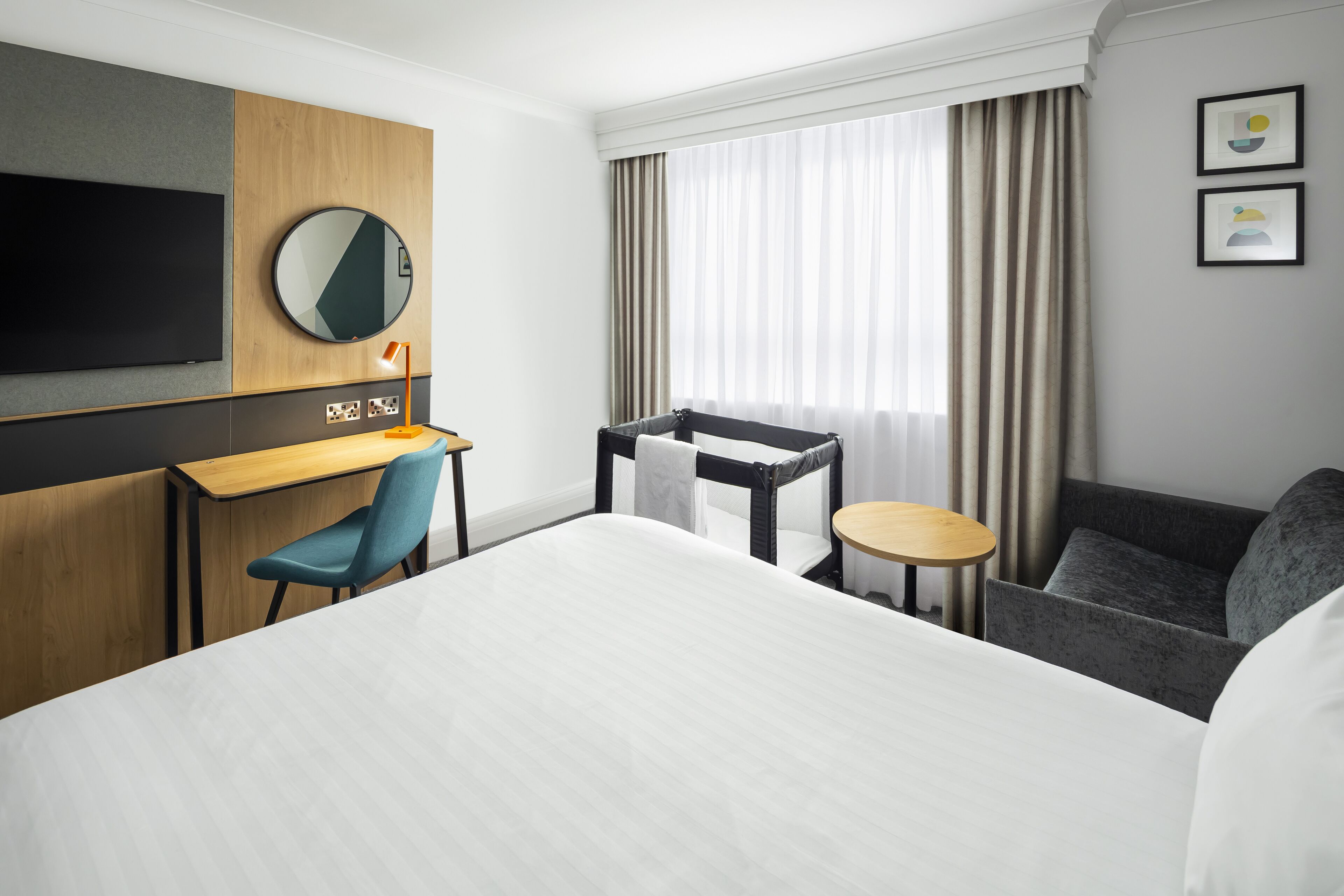 A hotel room with a large bed, a desk with a chair, a small table, a sofa, a TV mounted on the wall, and a window with curtains.