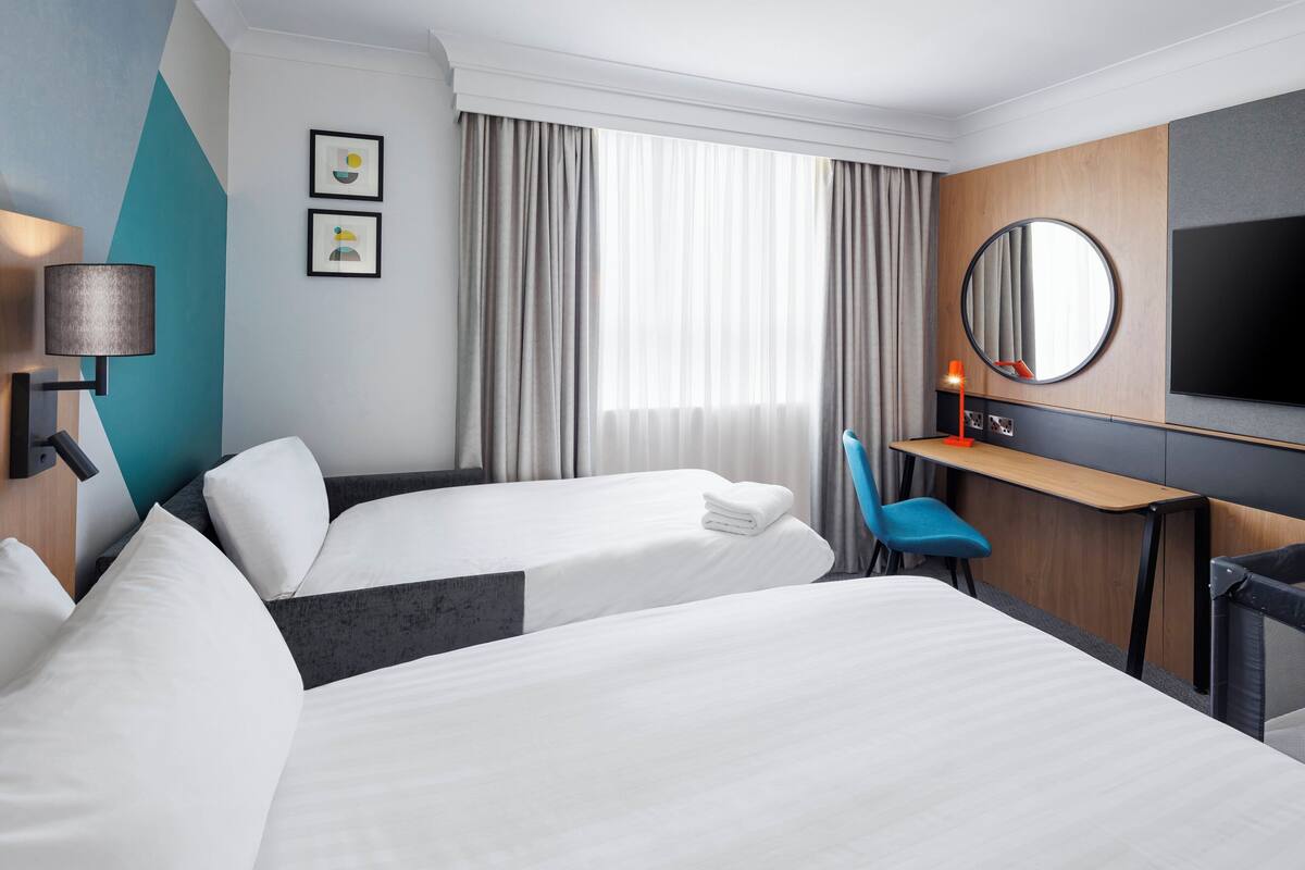 A hotel room with two beds, a desk, a chair, a TV, and a large window with curtains.