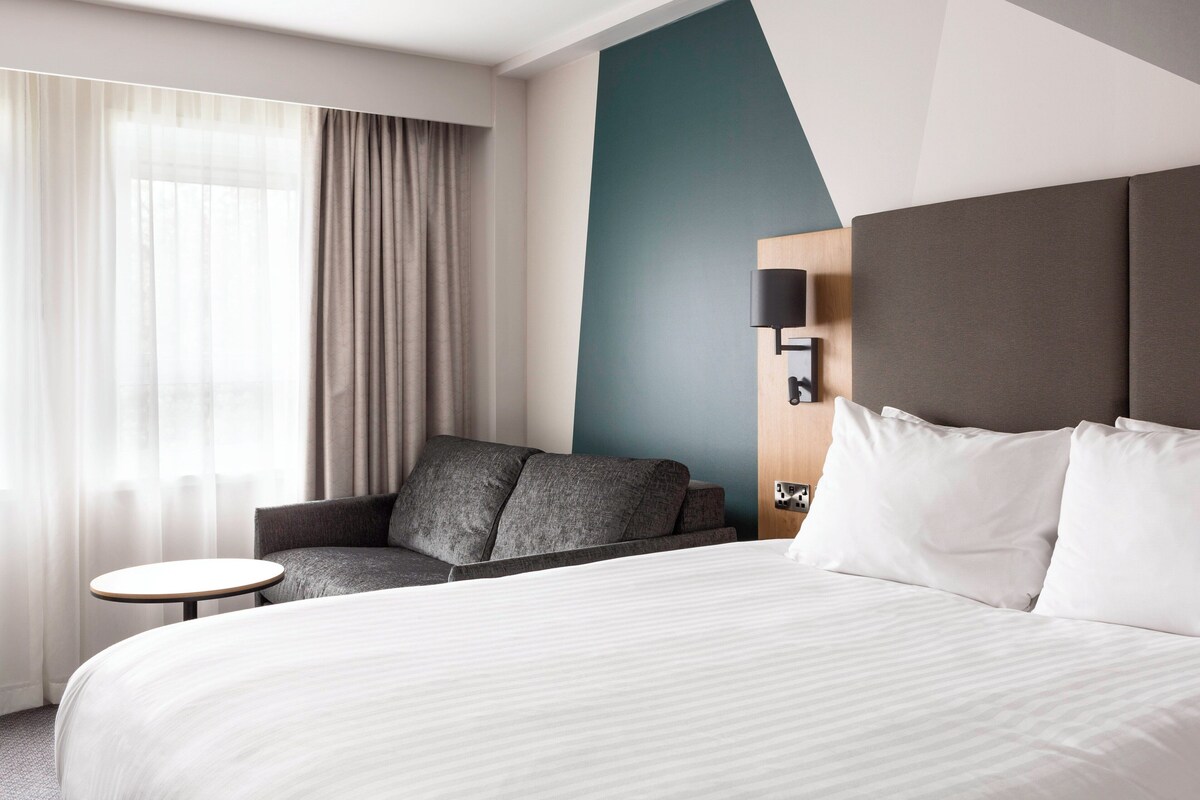 A hotel room with a bed, a sofa, a small table, and a wall-mounted lamp.
