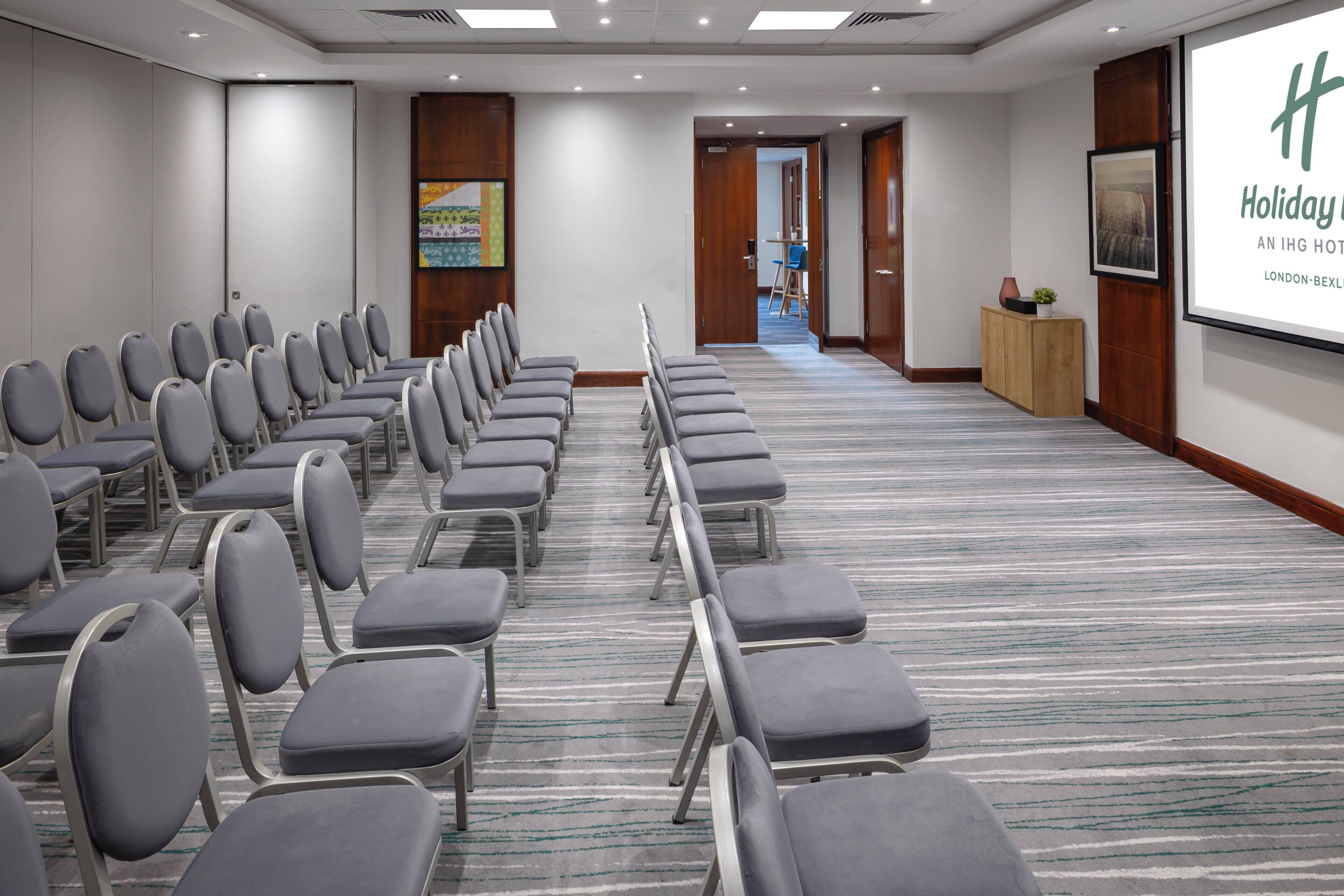 A conference room with rows of grey chairs, a carpeted floor, and a projection screen on the wall.
