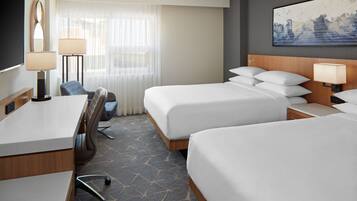 A hotel room with two beds, a desk, a chair, and a large window with sheer curtains.