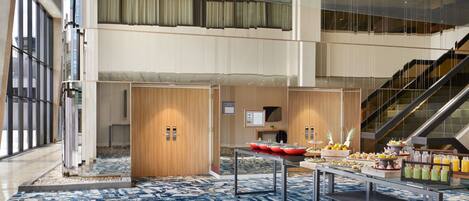 Un moderno vestíbulo de hotel con un amplio atrium, una escalera y una zona de buffet con diversas opciones de comida y bebida.