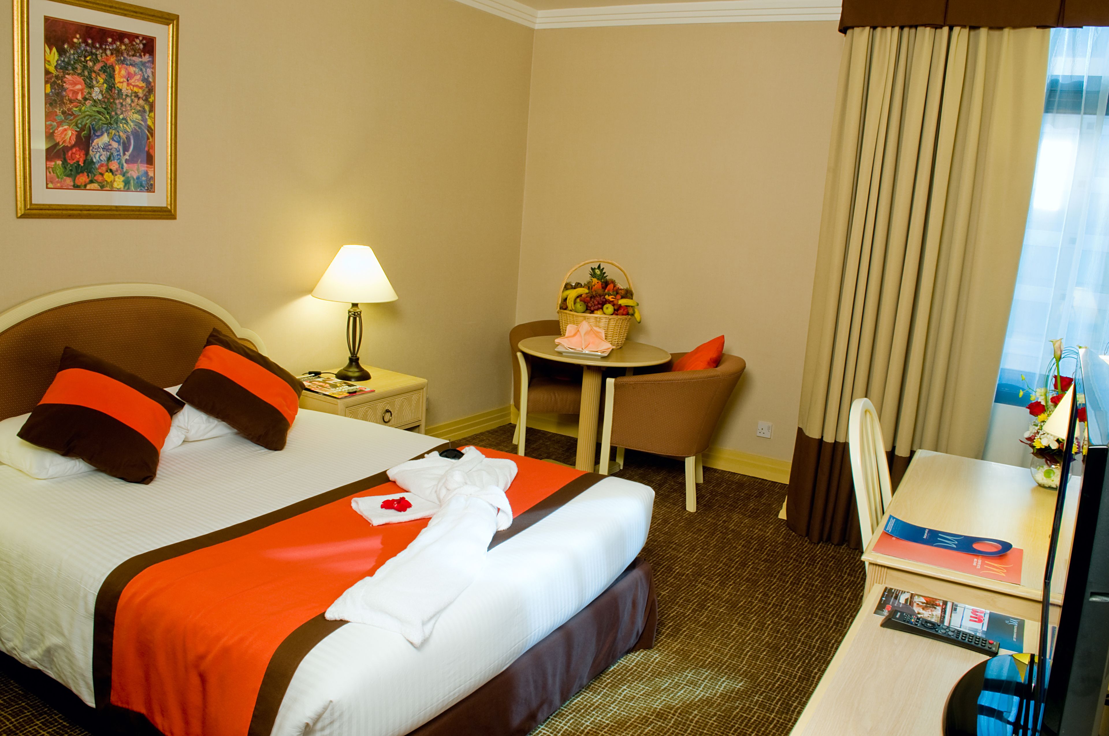 Habitación de hotel con cama, escritorio, silla, lámpara, mesita con cesta de fruta y un cuadro en la pared.