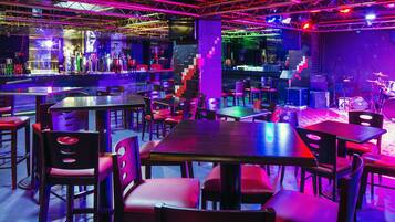 Zona de bar con mesas y sillas, un escenario con equipo musical y una barra con botellas.