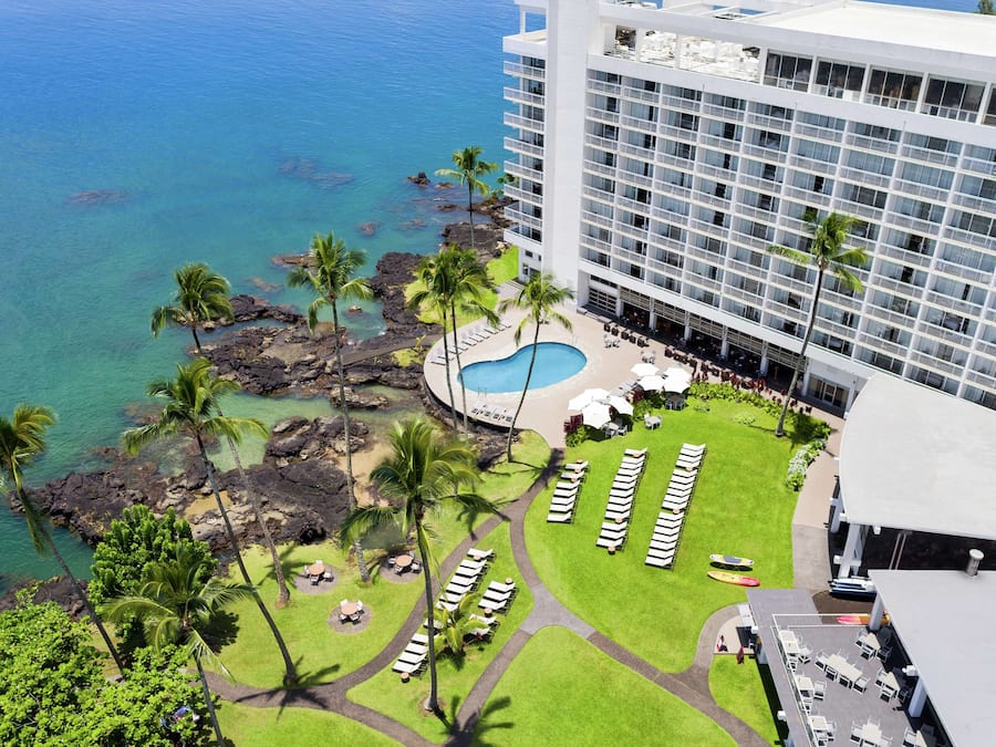 A hotel with a pool surrounded by palm trees and lounge chairs, overlooking a clear blue ocean.