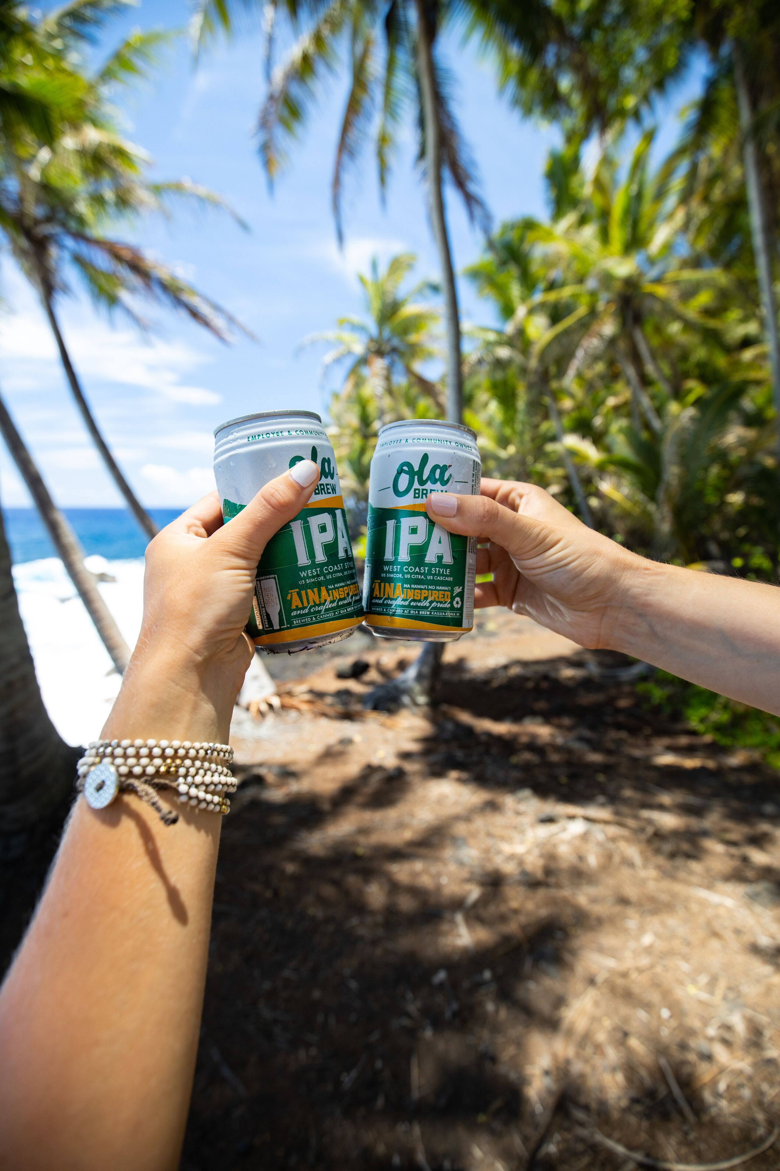 Due persone che tengono lattine di birra IPA 'Ola', con palme e una spiaggia sullo sfondo.